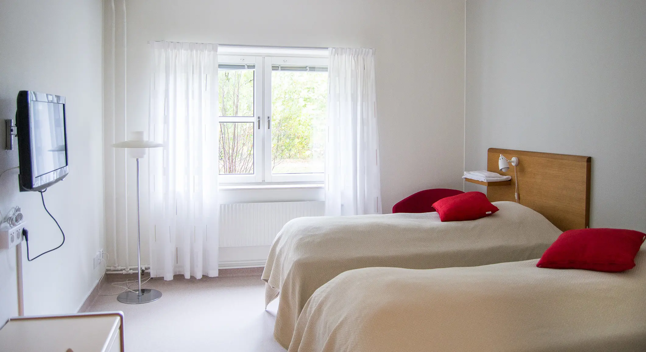 Ett rum där man ser två sängar som står bredvid varandra, nattduksbord, en TV som hänger på väggen och ett fönster med fin natur utanför.